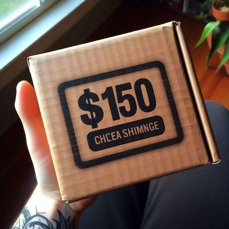 Hand holding a brown shipping box with $150 price label printed on top, indoor setting