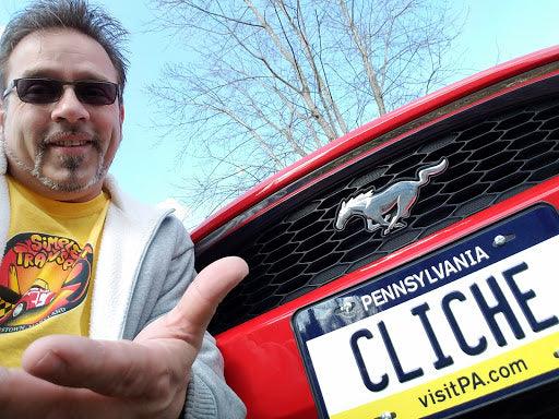 Man smiling next to red Ford Mustang with Pennsylvania novelty license plate reading CLICHE