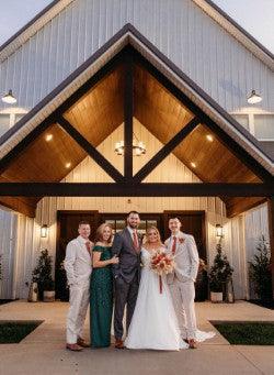 Wedding party posing in front of rustic barn venue at sunset, bride and groom centered